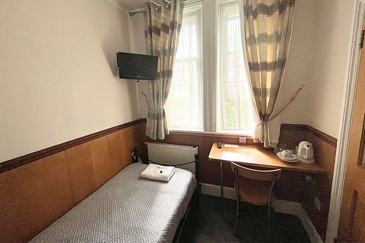 Egy kis, kompakt szoba, amelyben egyetlen ágy, egy íróasztal, egy szék, a falra szerelt televízió és egy függönnyel ellátott ablak található.