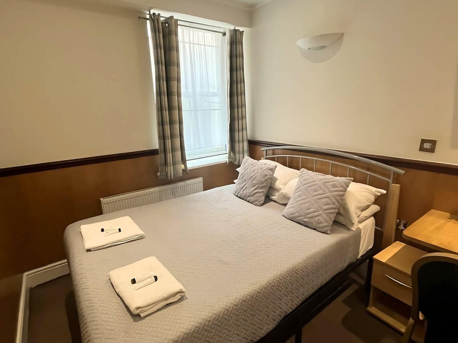 A hotel room with a bed, a bedside table, a chair, a window with curtains, and a wall-mounted light fixture.