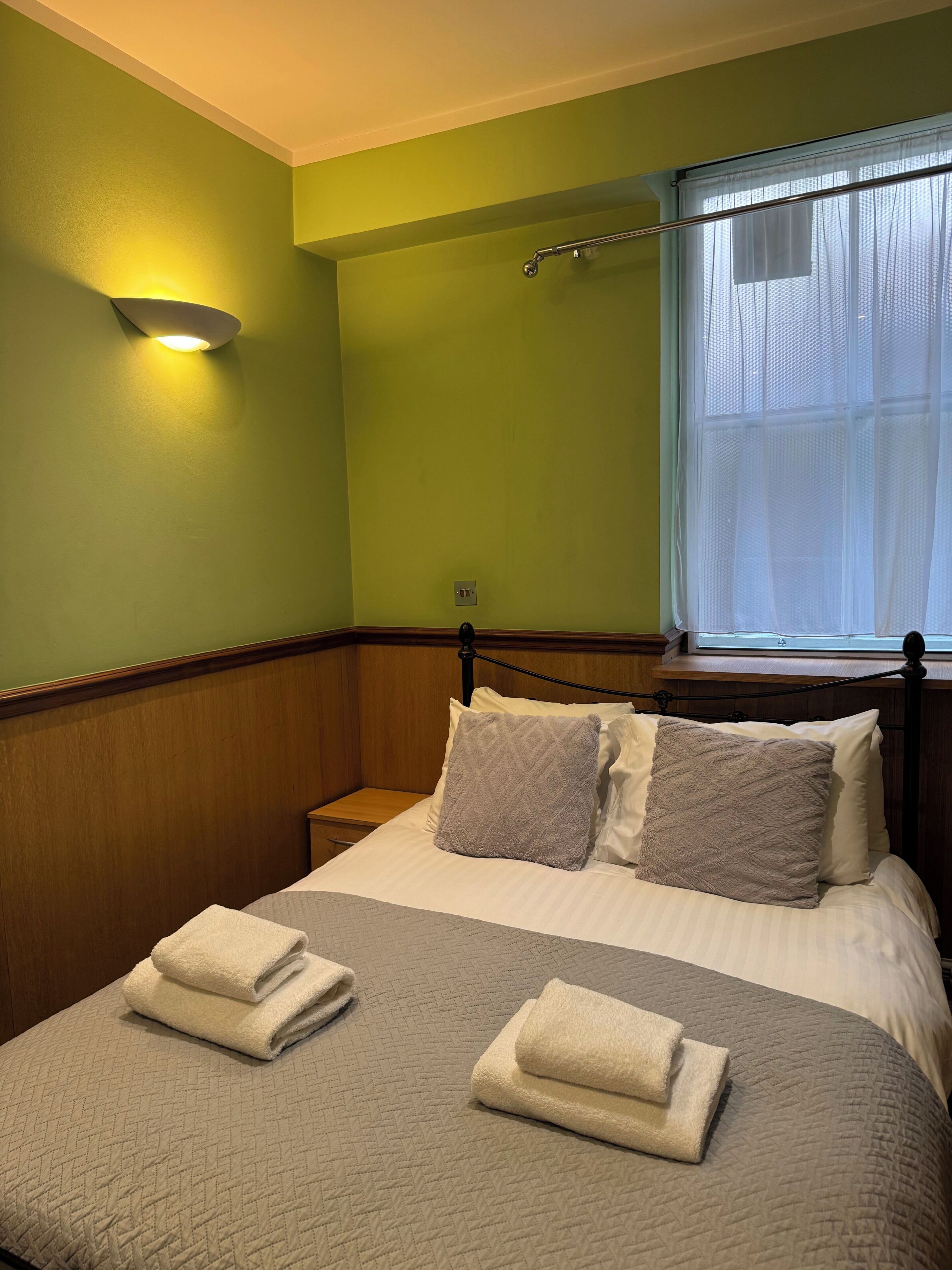 A neatly made bed with pillows and folded towels, a bedside table, a wall-mounted light, and a window with sheer curtains.