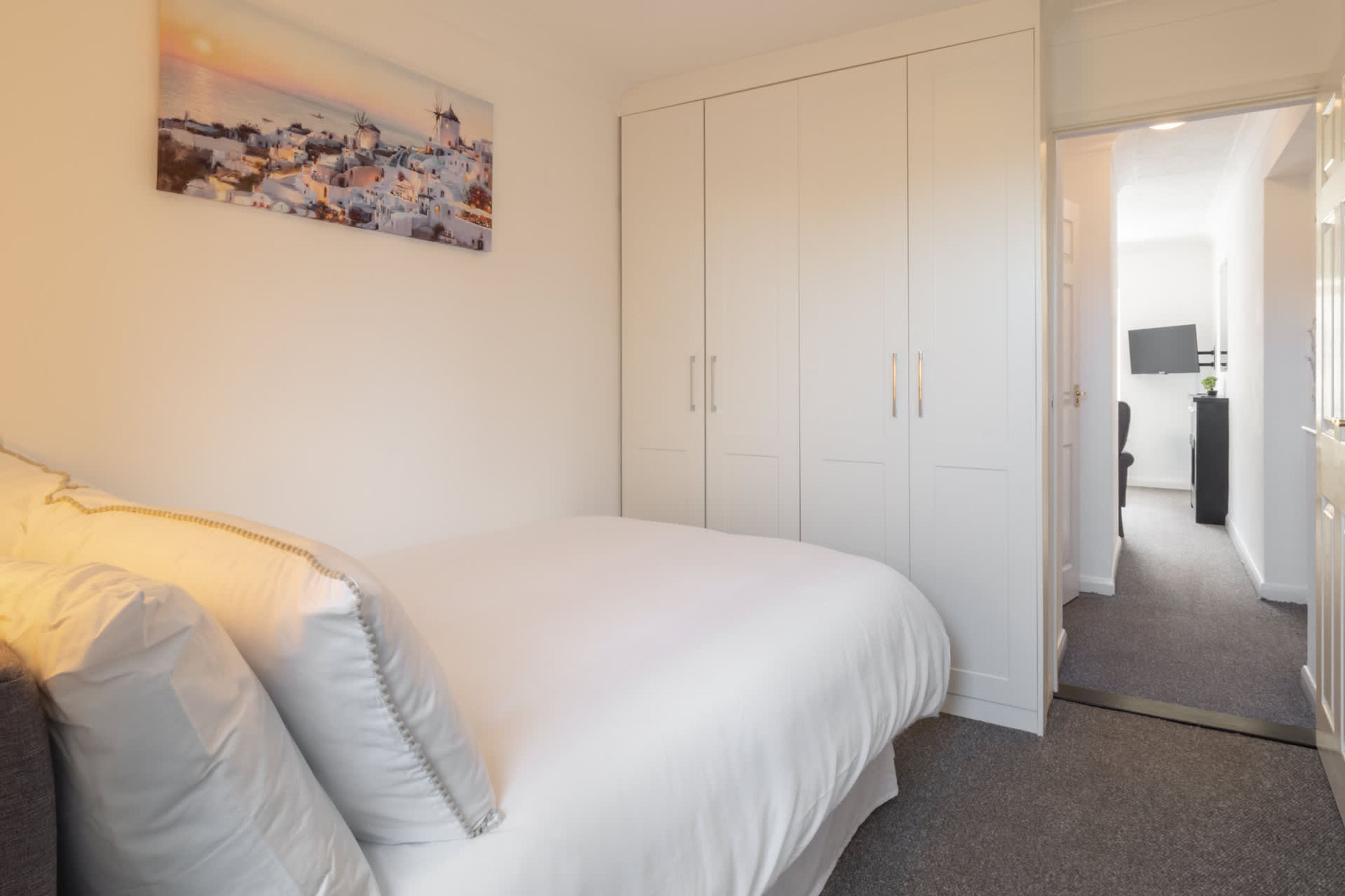 A bedroom with a bed, white wardrobe, and a painting of a coastal town.