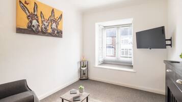 A modern living room with a grey sofa, a small table with a plant, a painting of donkeys, a window with blinds, and a mounted TV.