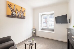 A modern living room with a grey sofa, a small table with a plant, a painting of donkeys, a window with blinds, and a mounted TV. - Host Stay St Martin s Road (Canterbury)