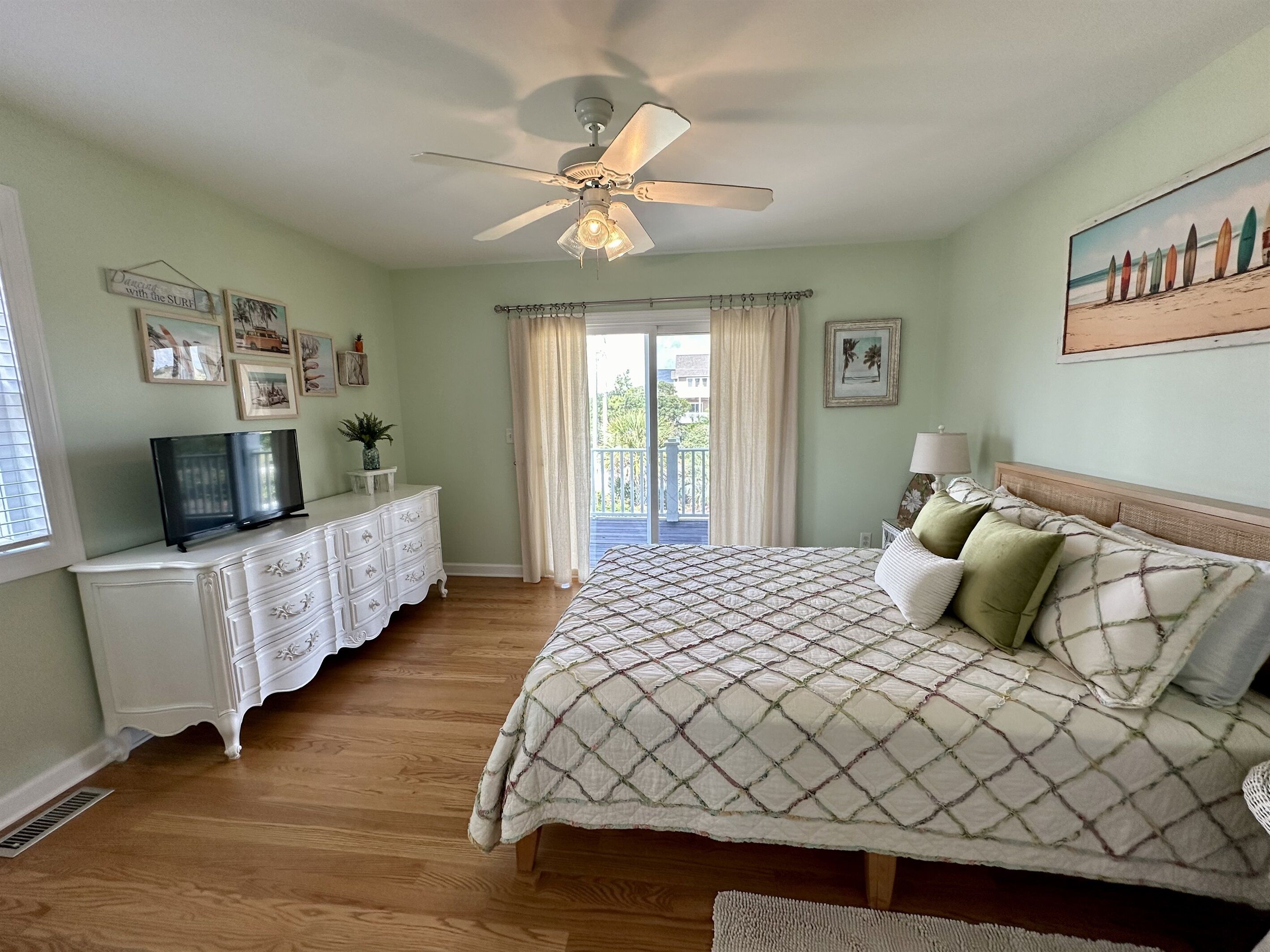 A bedroom with a bed, dresser, television, ceiling fan, and artwork on the walls.