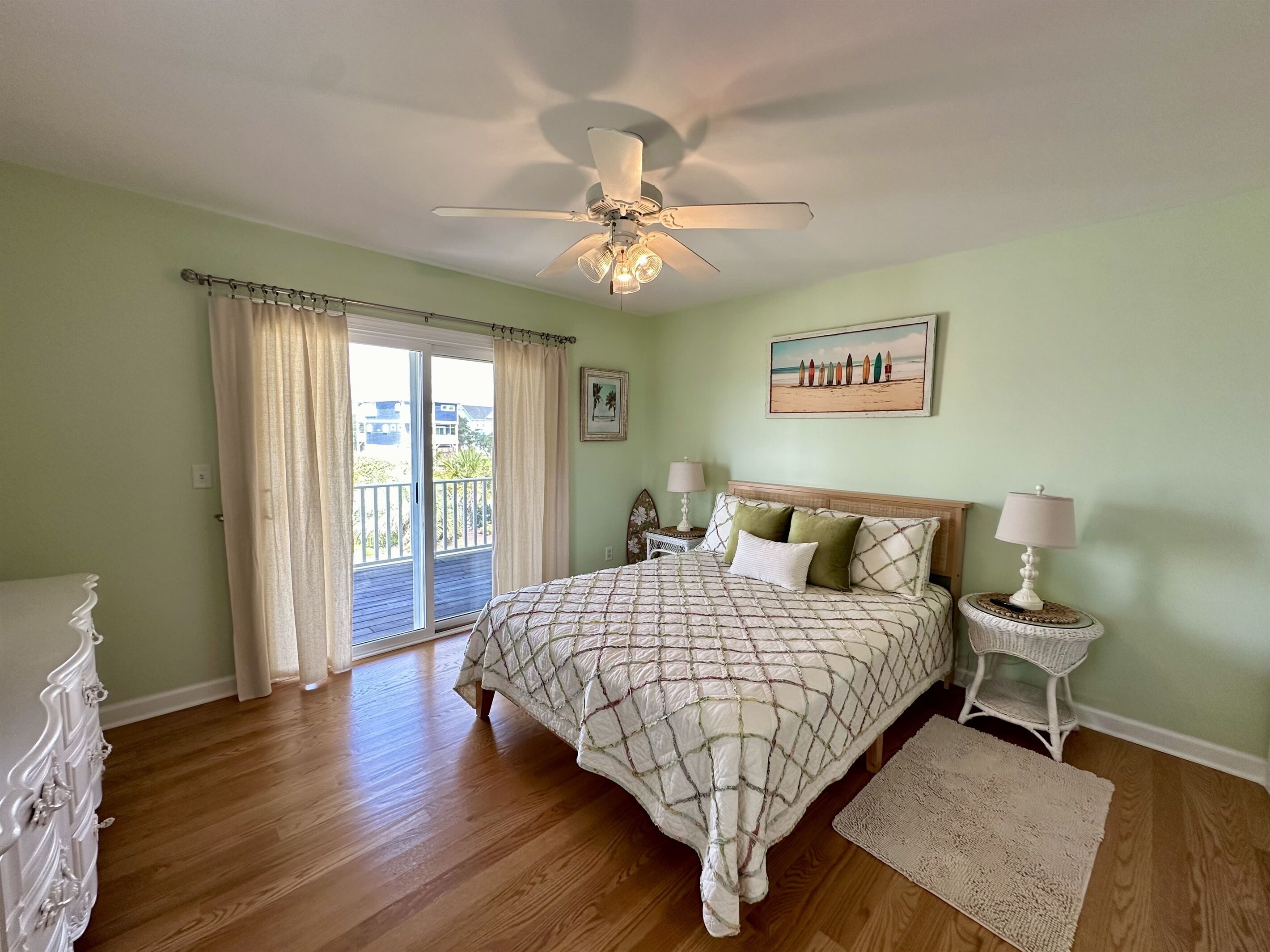 Une chambre à coucher avec un lit, un ventilateur de plafond, une table de chevet, une lampe, un tableau et une vue sur l’extérieur, visible à travers une porte-fenêtre coulissante.