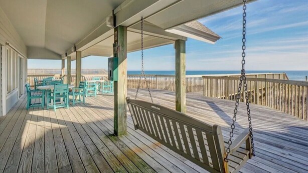 Una terraza de madera con una silla colgante y una mesa con sillas, con vista a una playa.