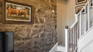 A stone wall with a framed picture of cattle, a flat-screen TV on a wooden stand, and a staircase with white railings. - 1 bedroom accommodation in Slaidburn (Slaidburn)