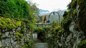A narrow stone pathway leading to a building with a metal archway, surrounded by greenery and a mountainous backdrop.
