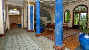 A grand hallway with blue columns, patterned floor tiles, and a view of a dining area.