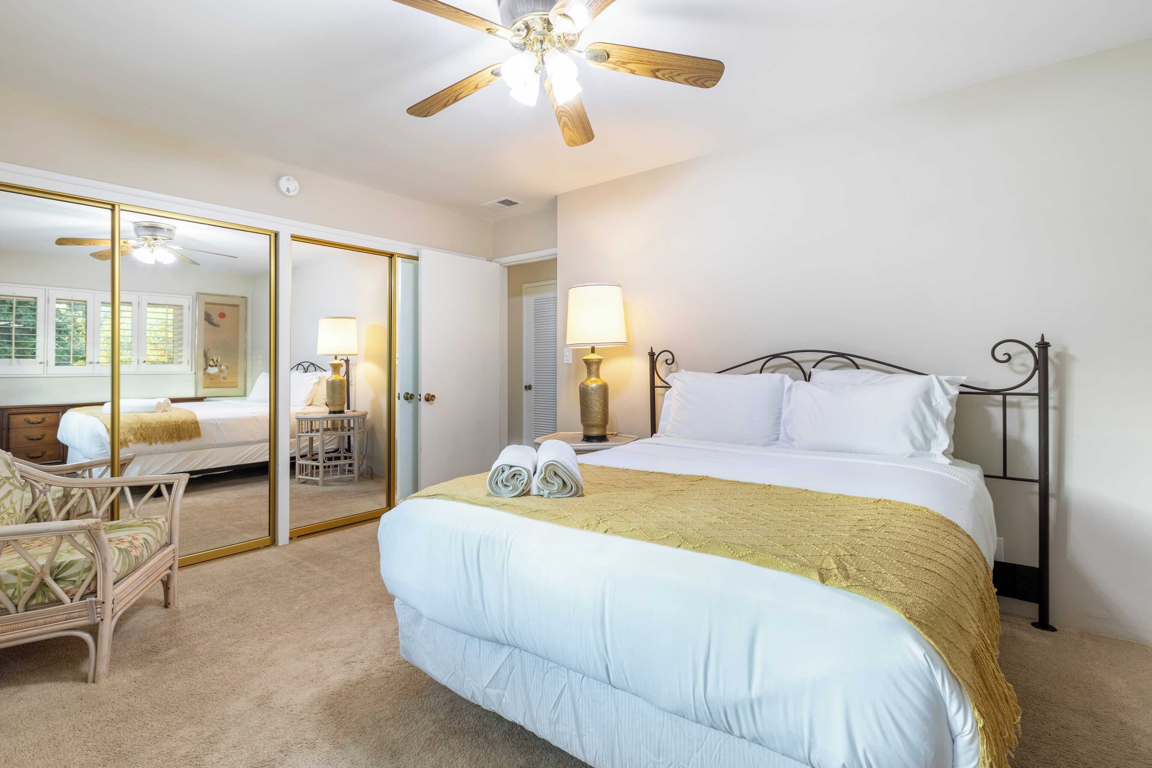 A hotel room with a large bed, a ceiling fan, a mirror, a chair, and a nightstand with a lamp.