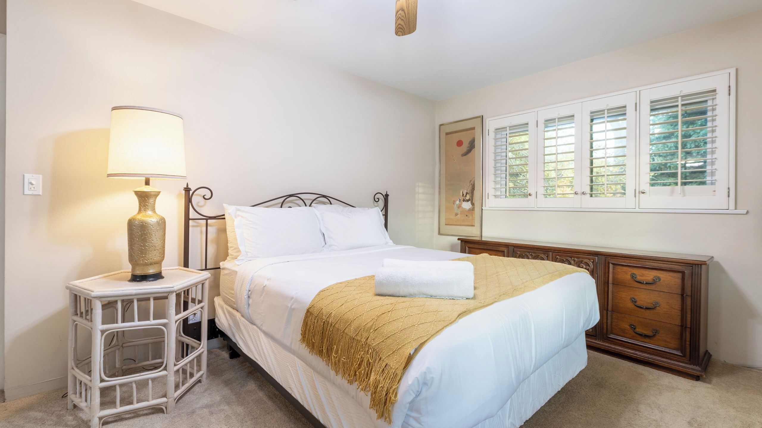A bedroom with a bed, bedside table, lamp, dresser, and window blinds.