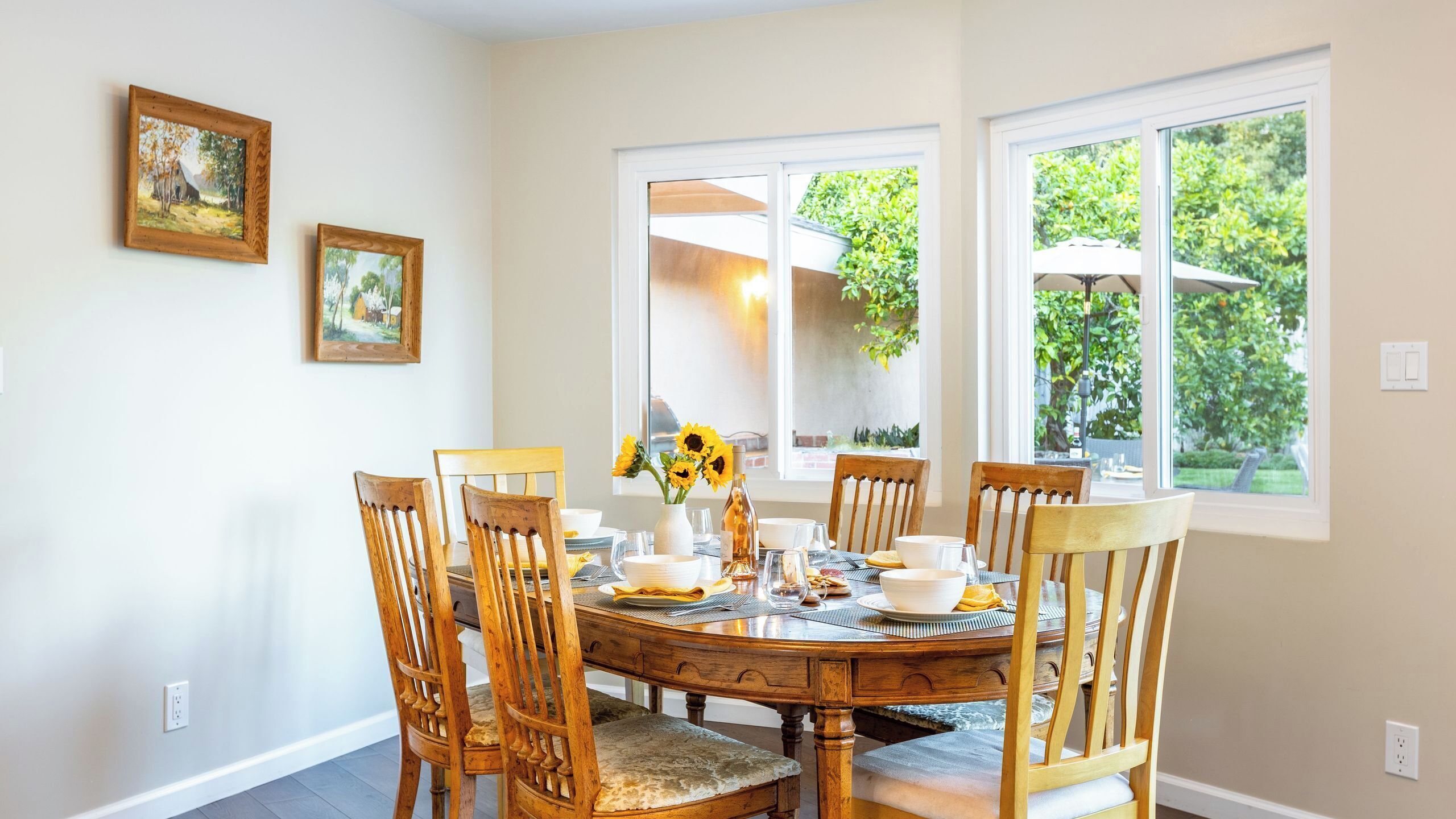 Uma sala de jantar com uma mesa de madeira redonda, seis cadeiras de madeira e duas pinturas emolduradas na parede.