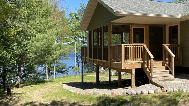 A wooden cabin with a deck, located near a body of water with trees in the background. - Long Lake Escape Three Lakes (Eagle River)