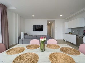 Uma sala de estar moderna com um sofá cinzento, uma televisão de tela plana, uma mesa de jantar com jogos americanos trançados e um vaso com flores.