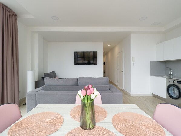 A modern living room with a grey sofa, a dining table with pink placemats, and a vase with flowers.