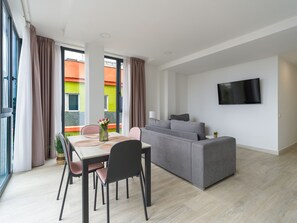 A modern living room with a grey sofa, a dining table with chairs, a flat-screen TV mounted on the wall, and large windows with curtains.