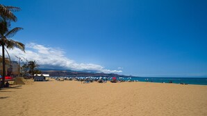 A wide sandy beach with umbrellas and beach chairs, palm trees, and a clear blue sky.