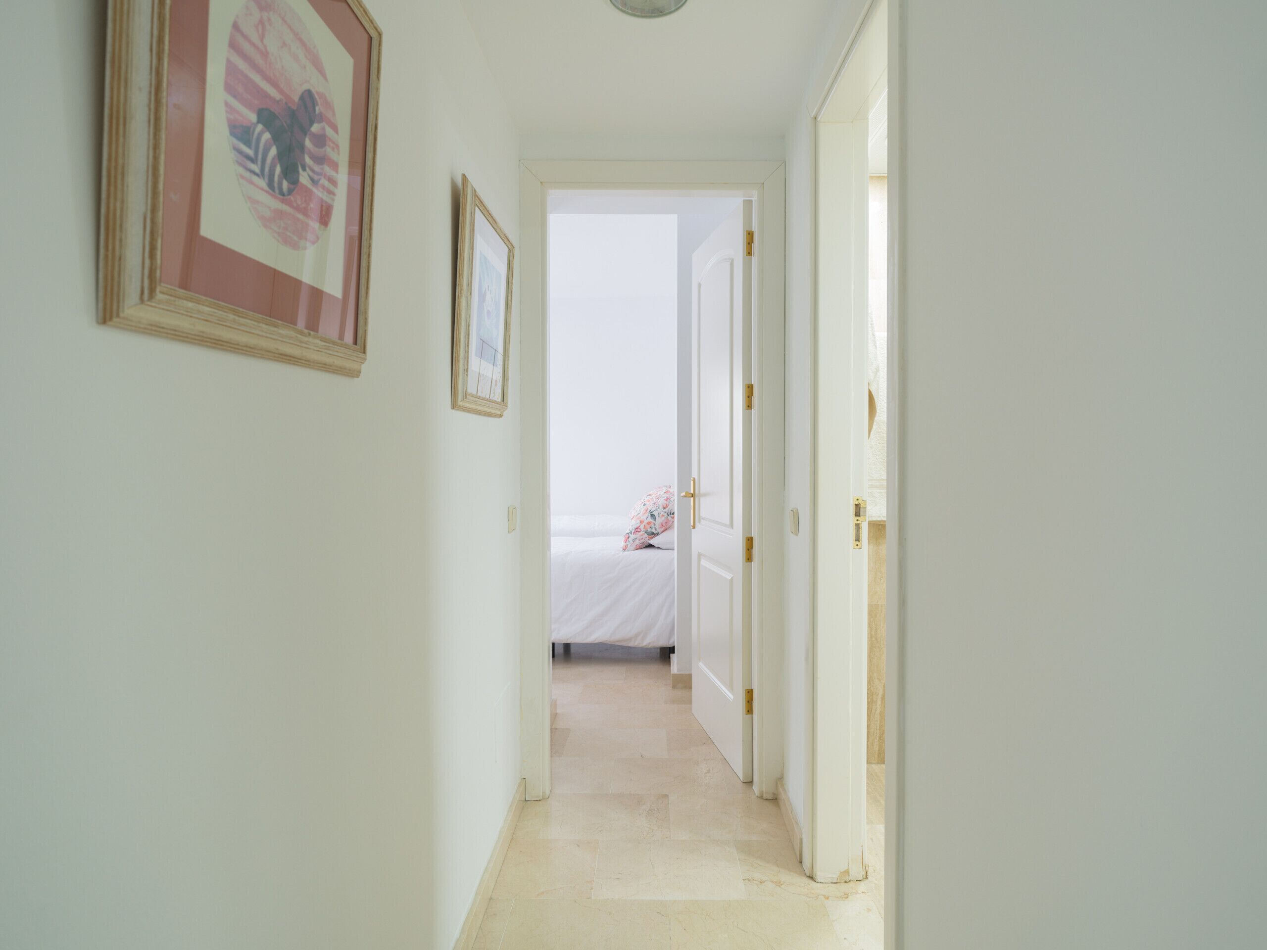 Un couloir étroit aux murs blancs, avec un tableau encadré sur le côté gauche, et un lit avec un coussin à motifs floraux au fond.
