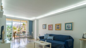 Un soggiorno con un divano blu, un tavolino bianco e un'area pranzo con tavolo e sedie in vetro. Quattro quadri sono appesi al muro.