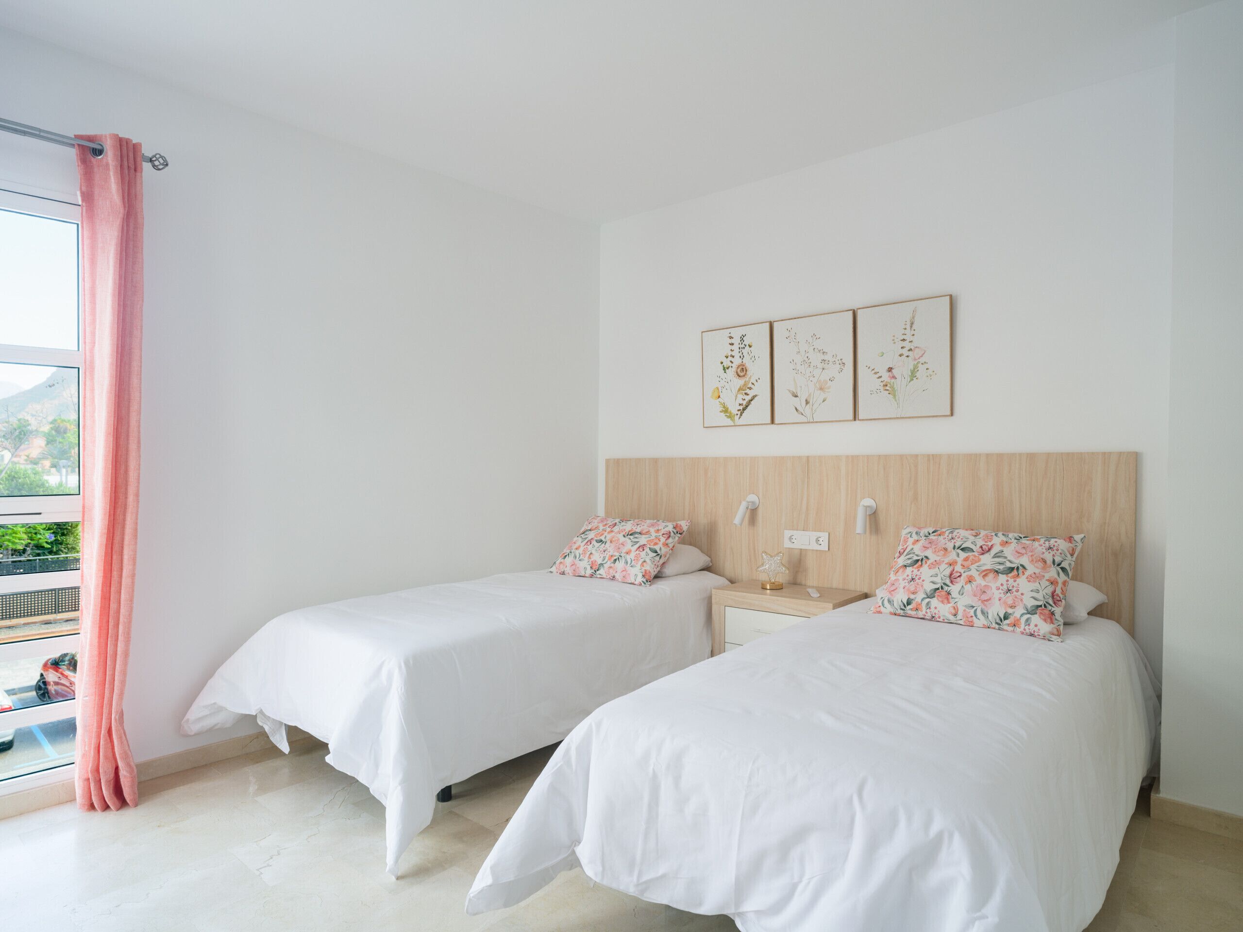 Ein Schlafzimmer mit zwei Betten, einem Fenster mit rosa Vorhang und drei floralen Bildern an der Wand.
