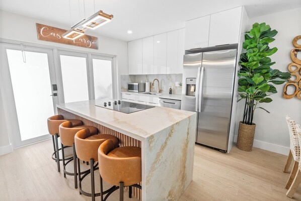 A modern kitchen with a central island, bar stools, stainless steel appliances, and a large potted plant.