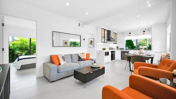 Ein modernes Wohnzimmer mit einer Couch, einem Couchtisch und orangefarbenen Sesseln. Es gibt einen Essbereich mit Tisch und Stühlen sowie eine Küche mit Herd und Schränken.