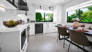 A modern kitchen with white cabinetry, a large window with a view of greenery, and a dining area with chairs and a table set for a meal.