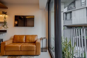 A modern living room with a leather sofa, a flat-screen TV, and a large window overlooking a balcony. - Vossa Bossa Agisse (São Paulo)
