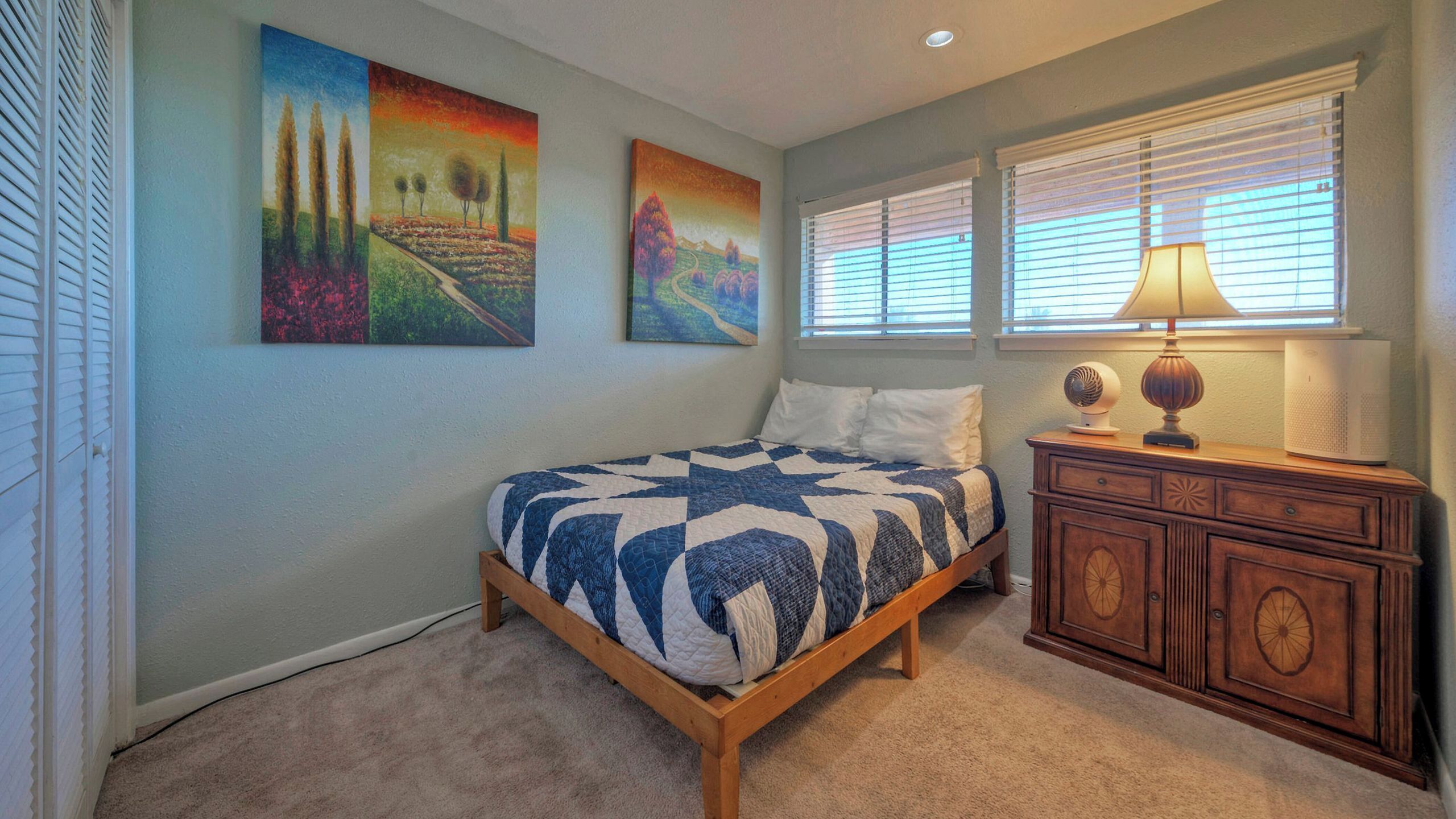 A bedroom with a bed, a dresser, a lamp, and three paintings on the wall.