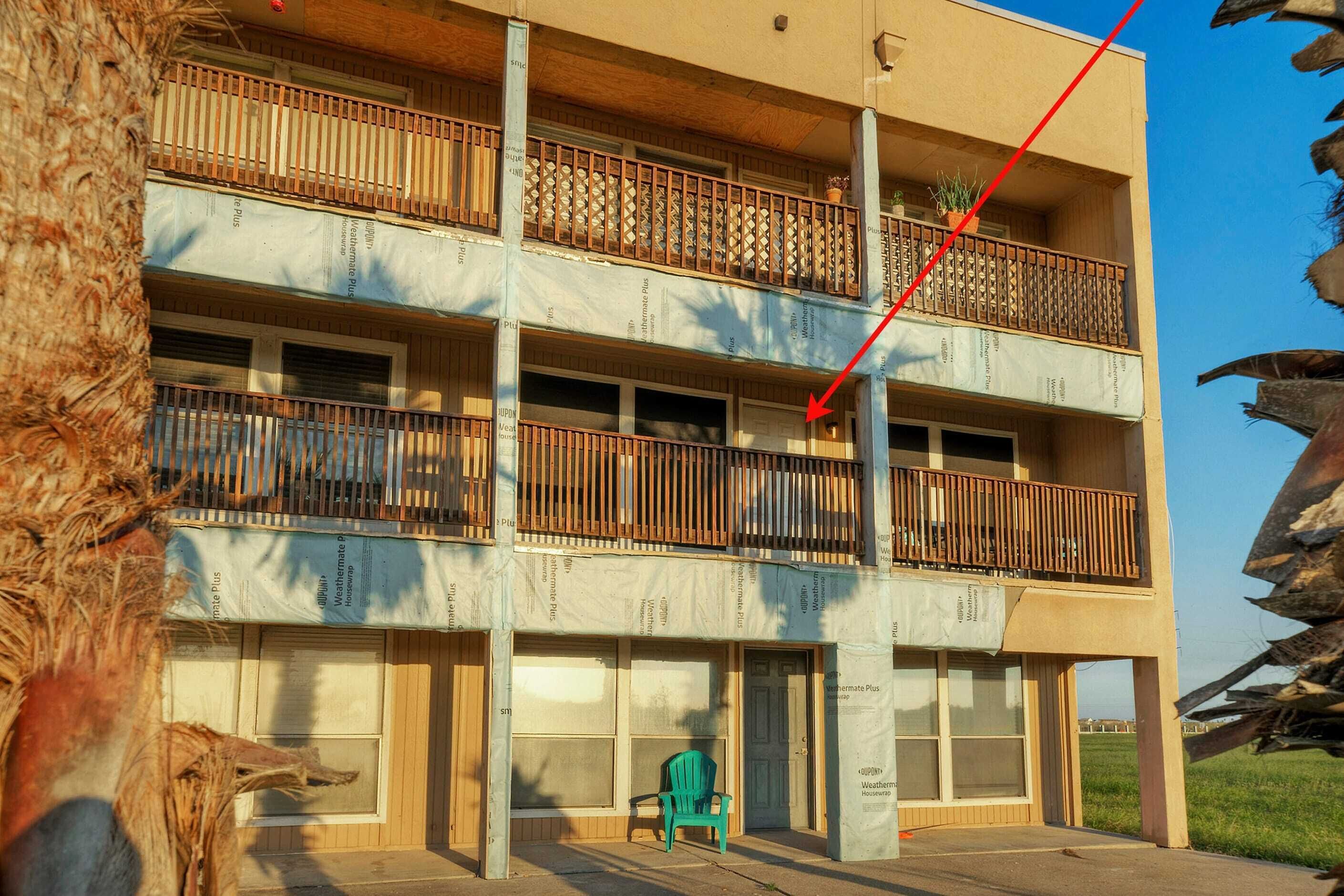 A multi-story building with balconies, a green chair outside, and a tree in the foreground.