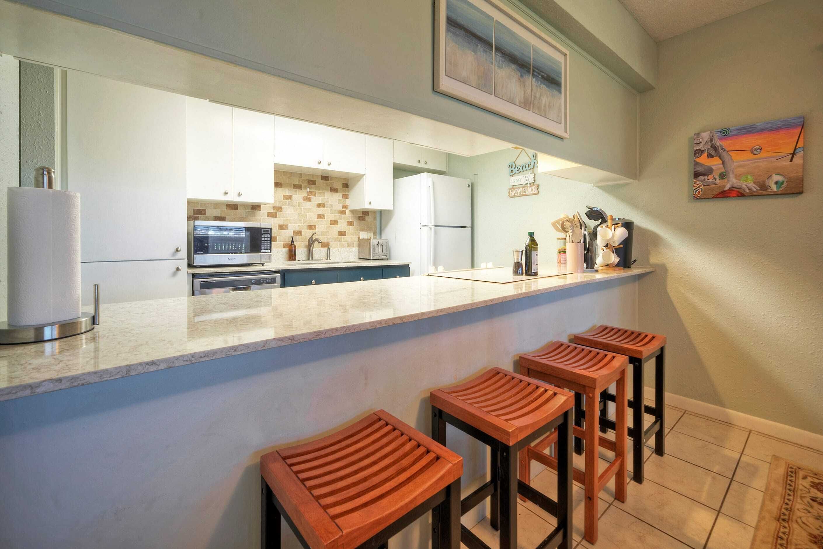 A kitchen with a marble countertop, white cabinets, a microwave, and a refrigerator. There are wooden stools at the counter.