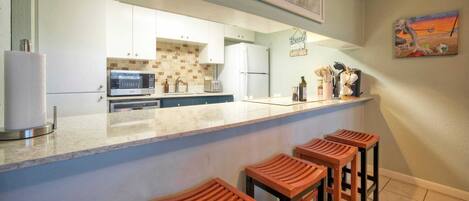 A kitchen with a marble countertop, white cabinets, a microwave, and a refrigerator. There are wooden stools at the counter.