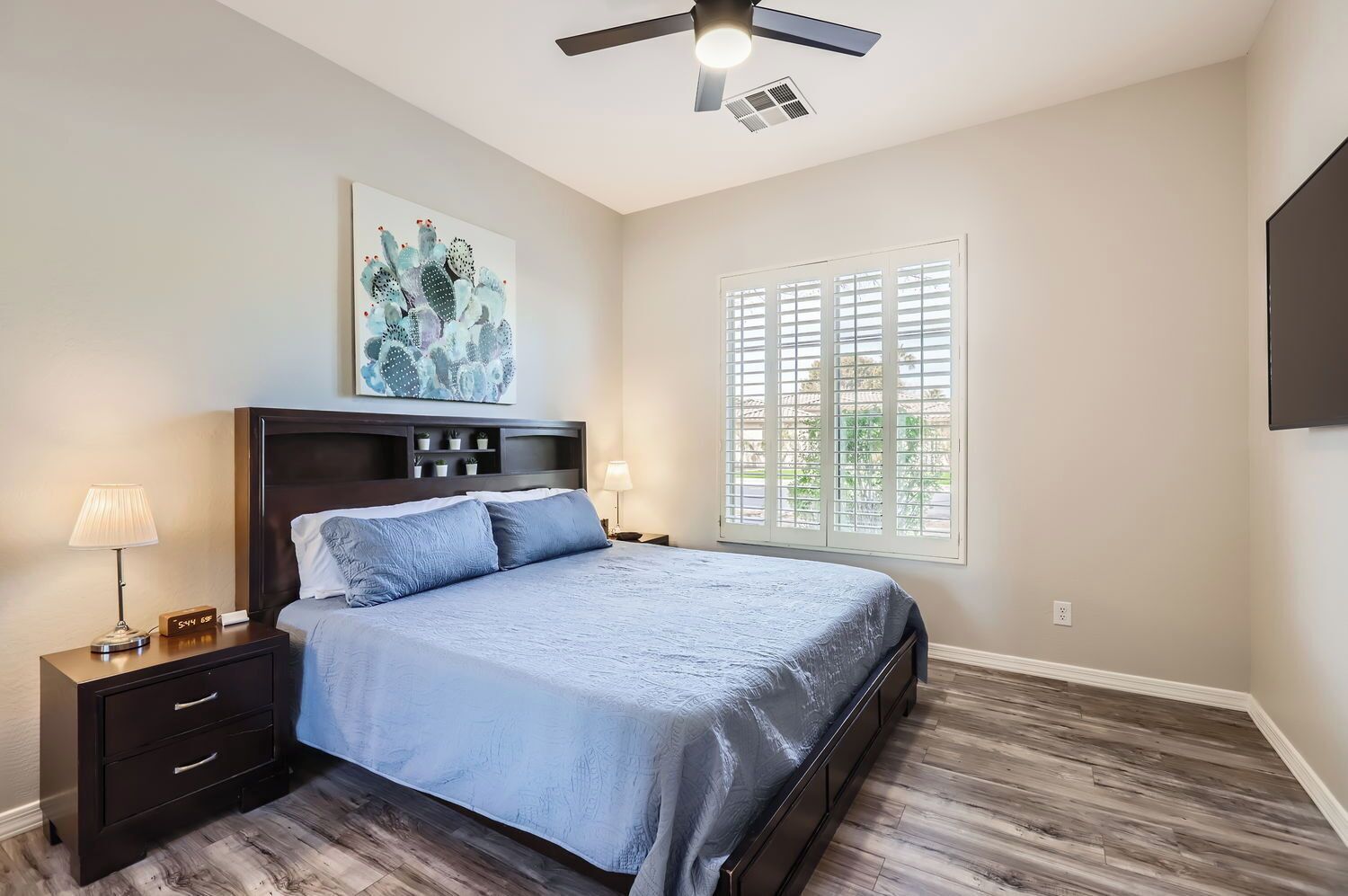 A bedroom with a bed, nightstands, a ceiling fan, a window with blinds, and a wall-mounted artwork.