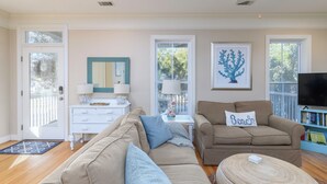 A living room with a beige sofa, a brown armchair, a coffee table, and a TV. There is a bookshelf with books and a framed picture on the wall. - Screened Porches | Ping-Pong Table Pet-Friendly | Walk to Beach & Pier (Tybee Island)