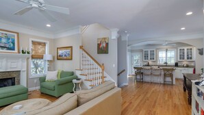 A spacious living room with a green sofa, a beige sofa, and a wooden staircase leading to a kitchen area. - Screened Porches | Ping-Pong Table Pet-Friendly | Walk to Beach & Pier (Tybee Island)