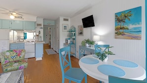 A compact kitchen with a dining area, a TV mounted on the wall, and a beach-themed painting. - Pools + Tennis | Walk to Shops & Restaurants | Coastal Condo (Tybee Island)