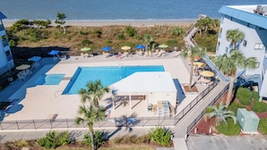 A pool area with lounge chairs and umbrellas, a building with a blue roof, and a view of the beach and ocean. - Pools + Tennis | Walk to Shops & Restaurants | Coastal Condo (Tybee Island)