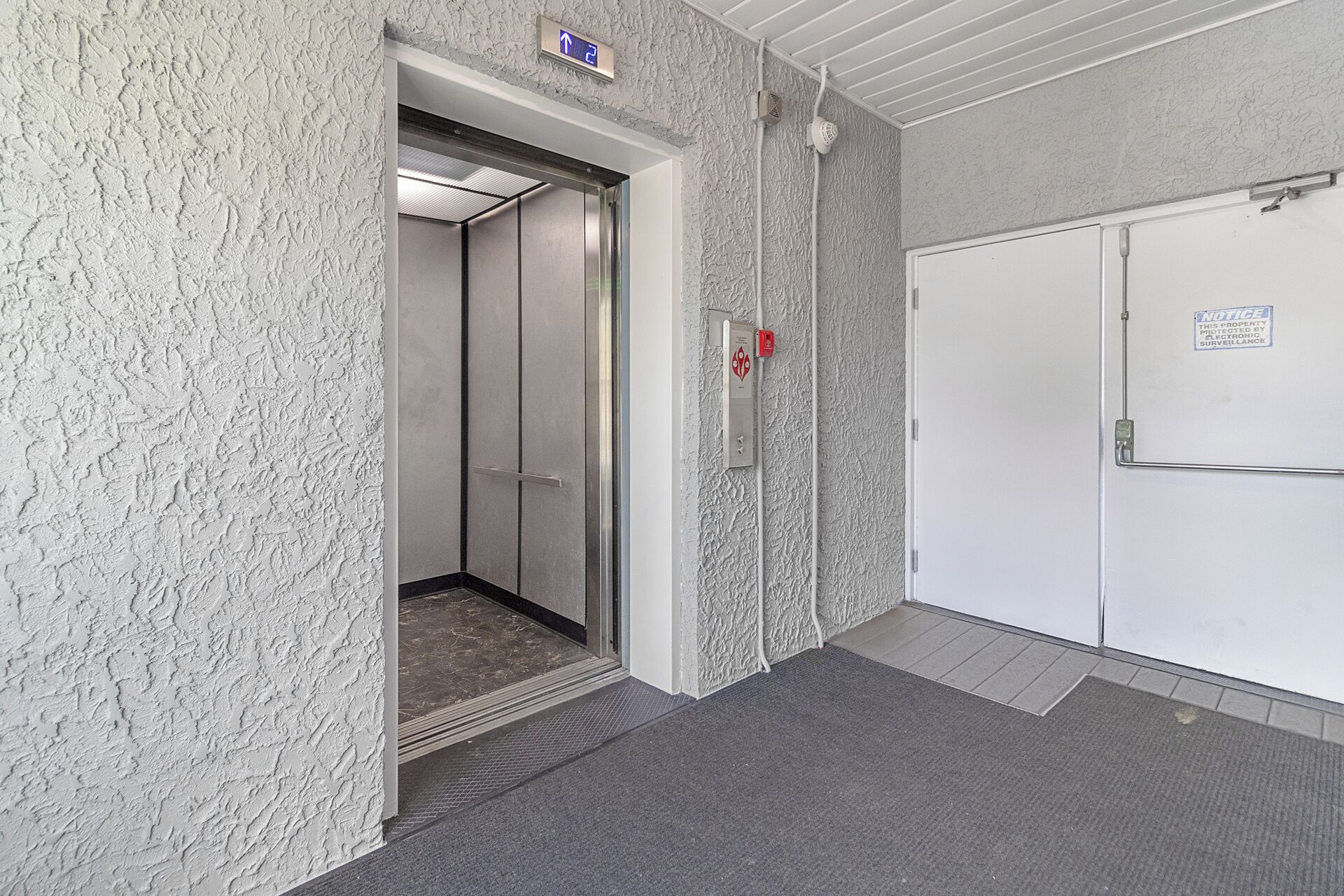 An elevator with a sign indicating the direction to the second floor, a fire alarm, and a security notice on the wall.