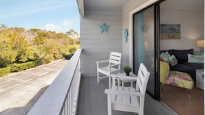 A balcony with white chairs and a small table, a sliding glass door leading to a living area with a sofa and cushions, and a star decoration on the wall. - Pools + Tennis | Walk to Shops & Restaurants | Coastal Condo (Tybee Island)