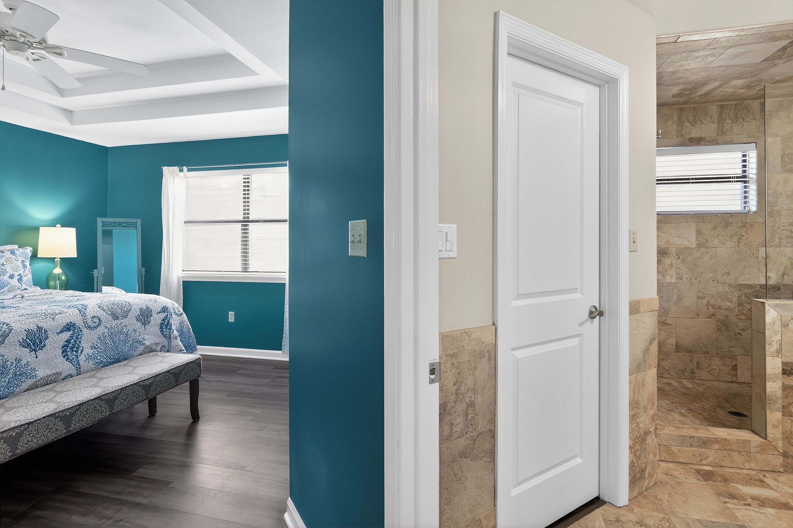 A bedroom with a bed, a bench, a window with blinds, and a door leading to a bathroom with a shower.