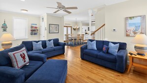 A living room with two blue sofas, a wooden floor, a ceiling fan, a dining area with a table and chairs, and a kitchen in the background.