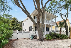 A two-story house with a porch, a white fence, and a tree in the foreground.