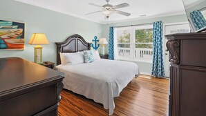 A bedroom with a bed, a ceiling fan, a window with curtains, a painting on the wall, and a nightstand with a lamp.