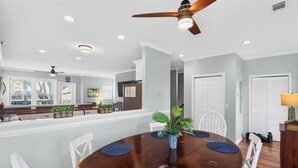 A dining area with a round table, white chairs, ceiling fans, recessed lighting, and a kitchen in the background.