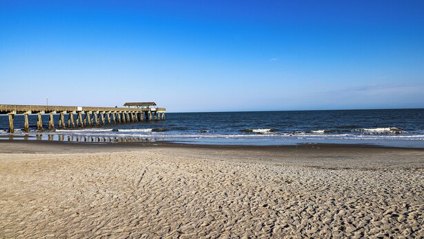A beach with a pier extending into the ocean. - Steps to the Beach | Ocean Glimpses from the Balcony (Tybee Island)