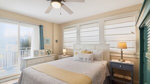 A bedroom with a bed, nightstands, a ceiling fan, a window with blinds, and a view of the outside. - Steps to the Beach | Ocean Glimpses from the Balcony (Tybee Island)