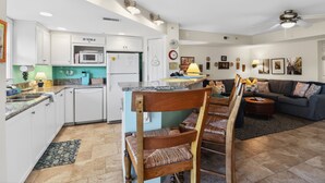 A modern kitchen with white cabinets, a microwave, and a refrigerator. There is a dining area with a wooden chair and a table. The living room has a dark-colored sofa and a coffee table. - Steps to the Beach | Ocean Glimpses from the Balcony (Tybee Island)