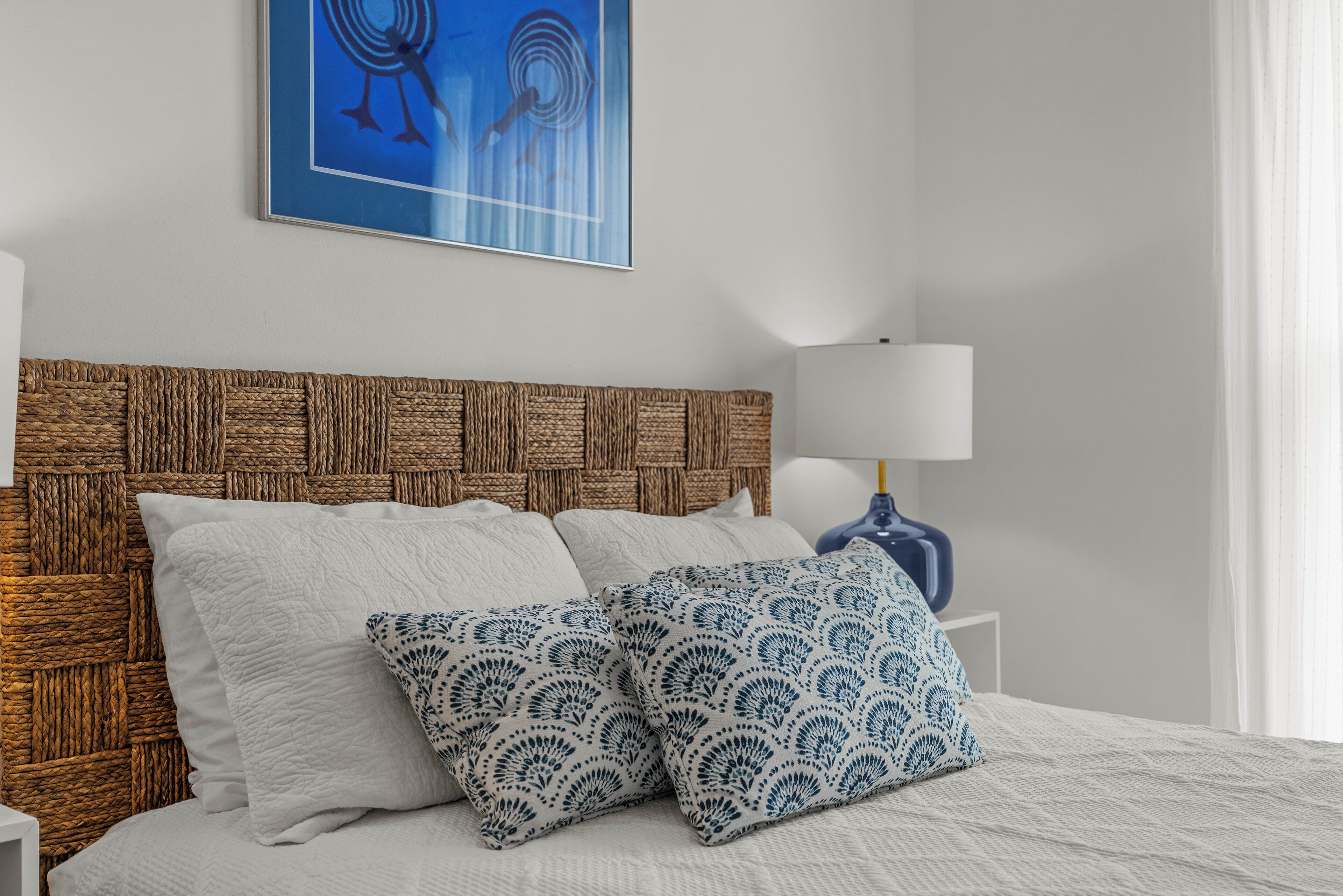 Un lit bien fait, avec des oreillers blancs et bleus à motifs, une tête de lit en osier et une œuvre d’art encadrée au-dessus.