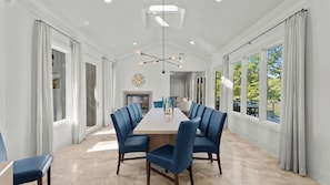 A spacious dining room with a long table, blue upholstered chairs, and large windows offering views of the outdoors.