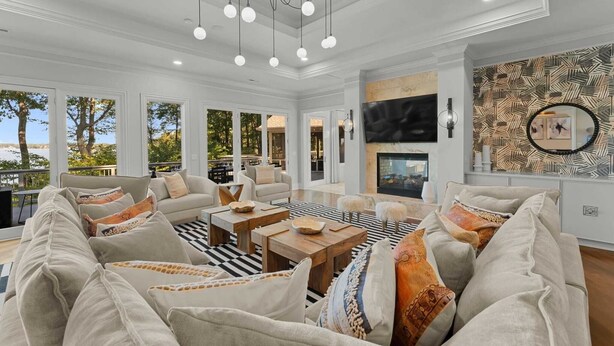 A spacious living room with a large sectional sofa, wooden coffee tables, and a chandelier. There are large windows with a view of trees and a balcony. - Modern Masterpiece by Avantstay Waterfront + Private Dock (Denver)