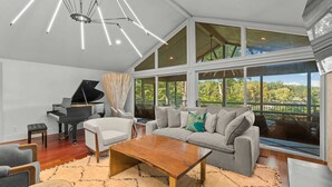 A spacious living room with a grand piano, a grey sofa with teal pillows, and a large window offering a view of greenery. - Modern Masterpiece by Avantstay Waterfront + Private Dock (Denver)